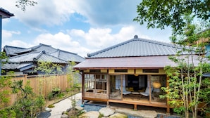 Une maison traditionnelle japonaise, avec une façade en bois, un toit recouvert de tuiles et un jardin agrémenté de rochers et de plantes.
