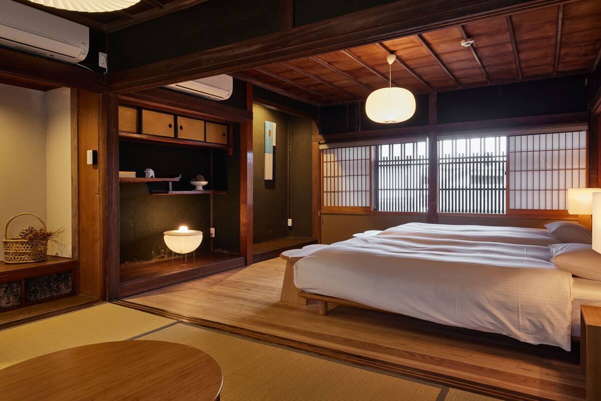 Ein traditioneller japanischer Raum mit Tatami-Boden, einem Bett mit weißer Bettwäsche, einer Hängeleuchte und Blick durch Shoji-Schirme.
