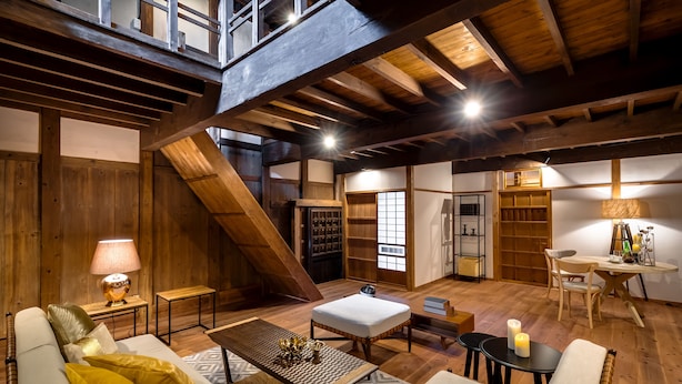 Un vaste espace de vie de style loft, doté d’un plafond haut, de poutres apparentes, d’un escalier intégré et d’un coin salon confortable avec un canapé, un pouf et une table basse.