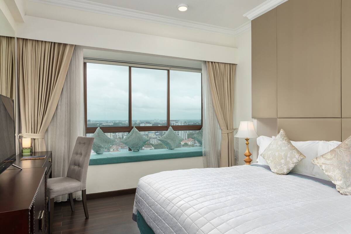 Une chambre d’hôtel avec un grand lit, un bureau, une chaise et une vue sur la ville par une grande fenêtre.