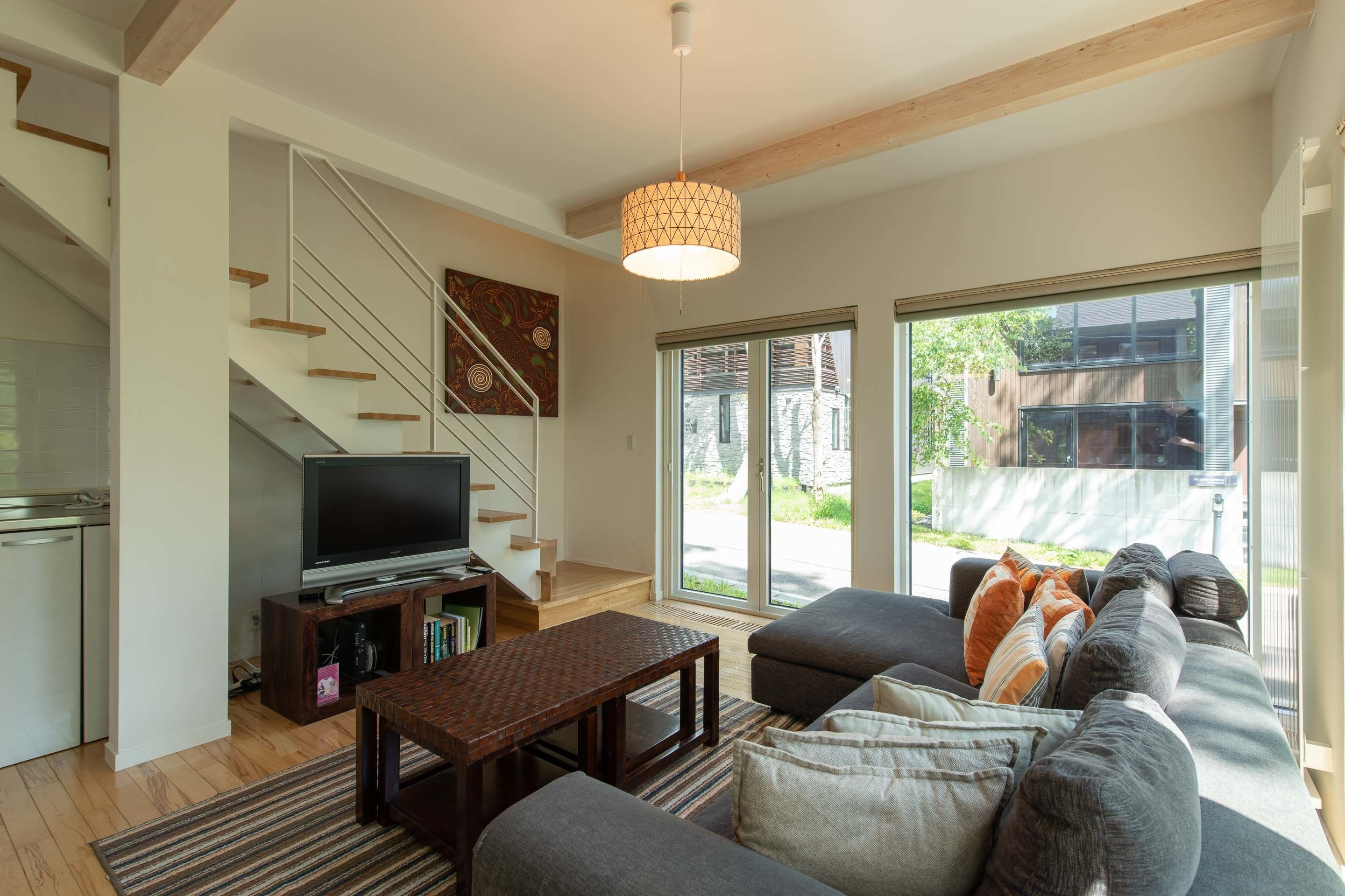 Ein modernes Wohnzimmer mit Treppe, Flachbildfernseher, Couchtisch aus Holz und grauem Sofa mit Kissen.