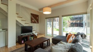 A modern living room with a staircase, a flat-screen TV, a wooden coffee table, and a gray sofa with cushions.
