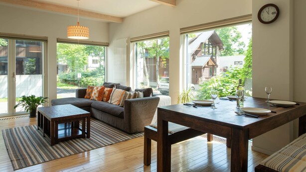A modern living room with a sofa, a dining table set for two, and large windows offering a view of the outdoors.