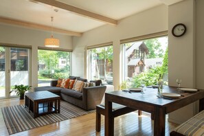 Una sala de estar moderna con un sofá, una mesa de comedor para dos personas y grandes ventanales con vistas al exterior.