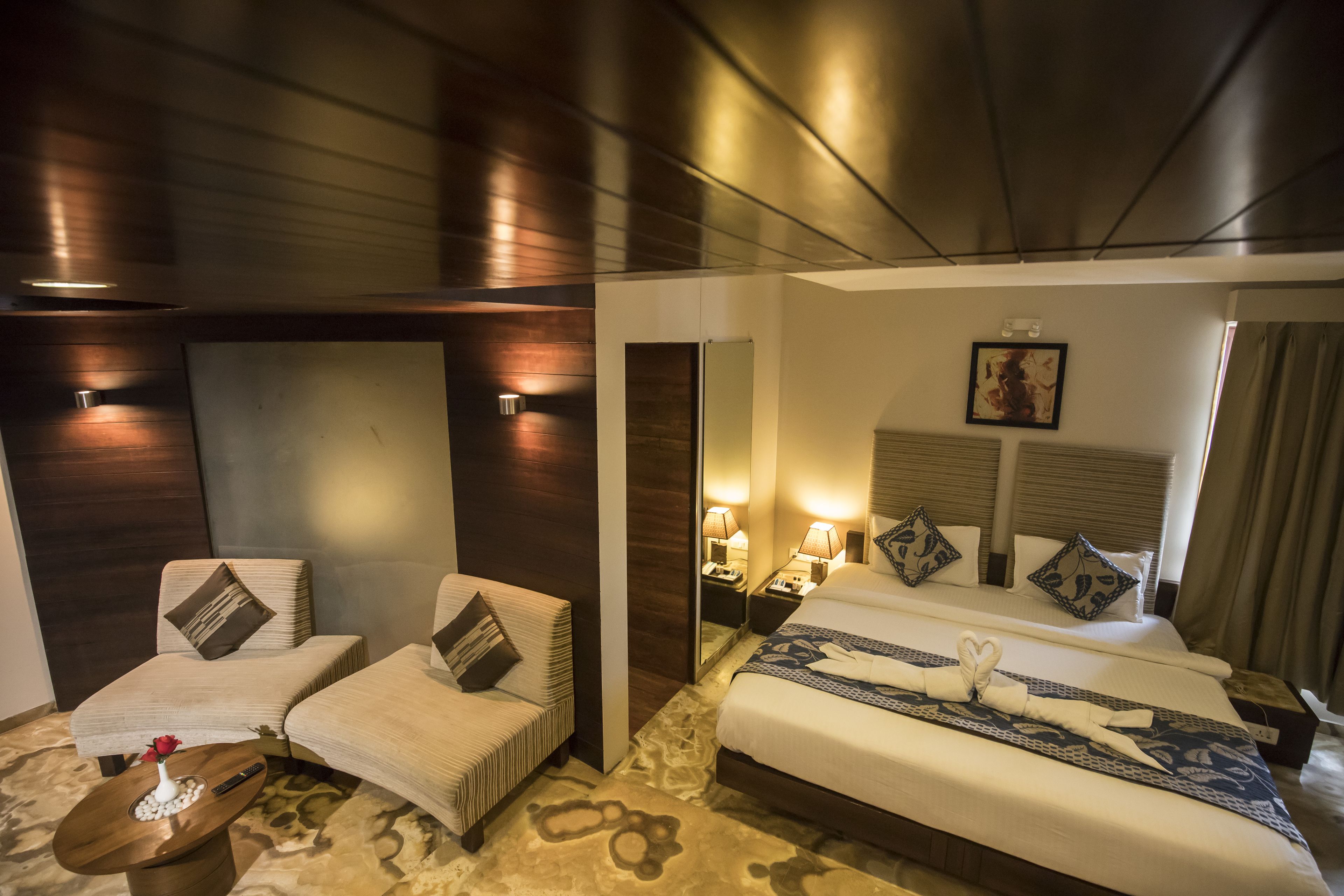 A hotel room with a bed, two armchairs, a small table with a vase, and a mirror.