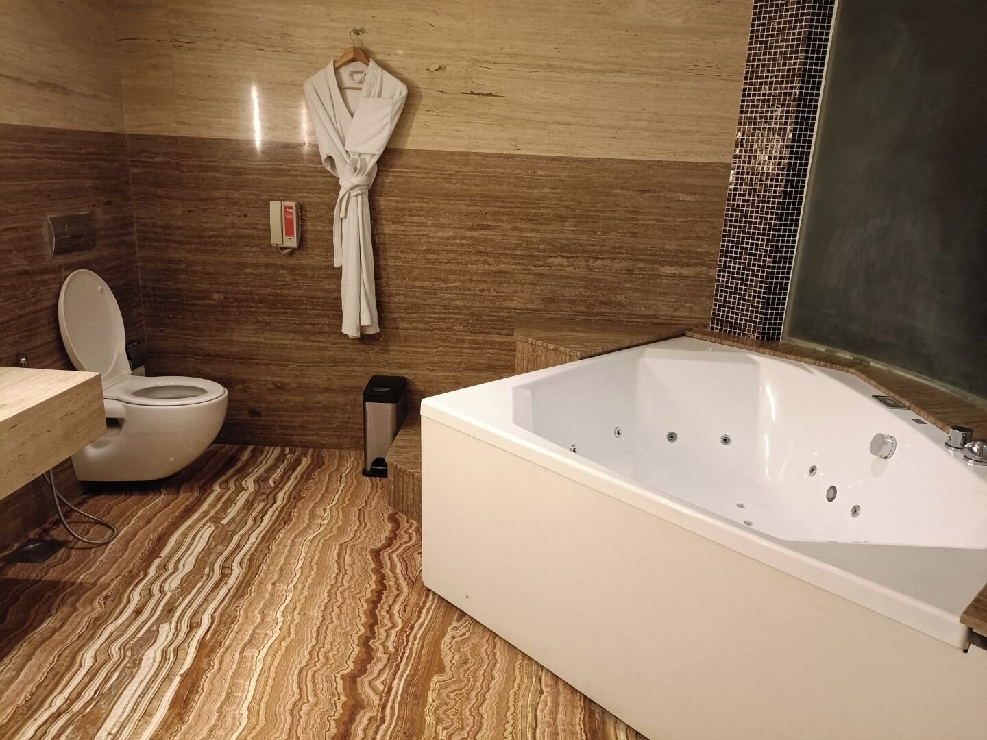 A modern bathroom with a marble floor, a white bathtub, a toilet, and a robe hanging on the wall.