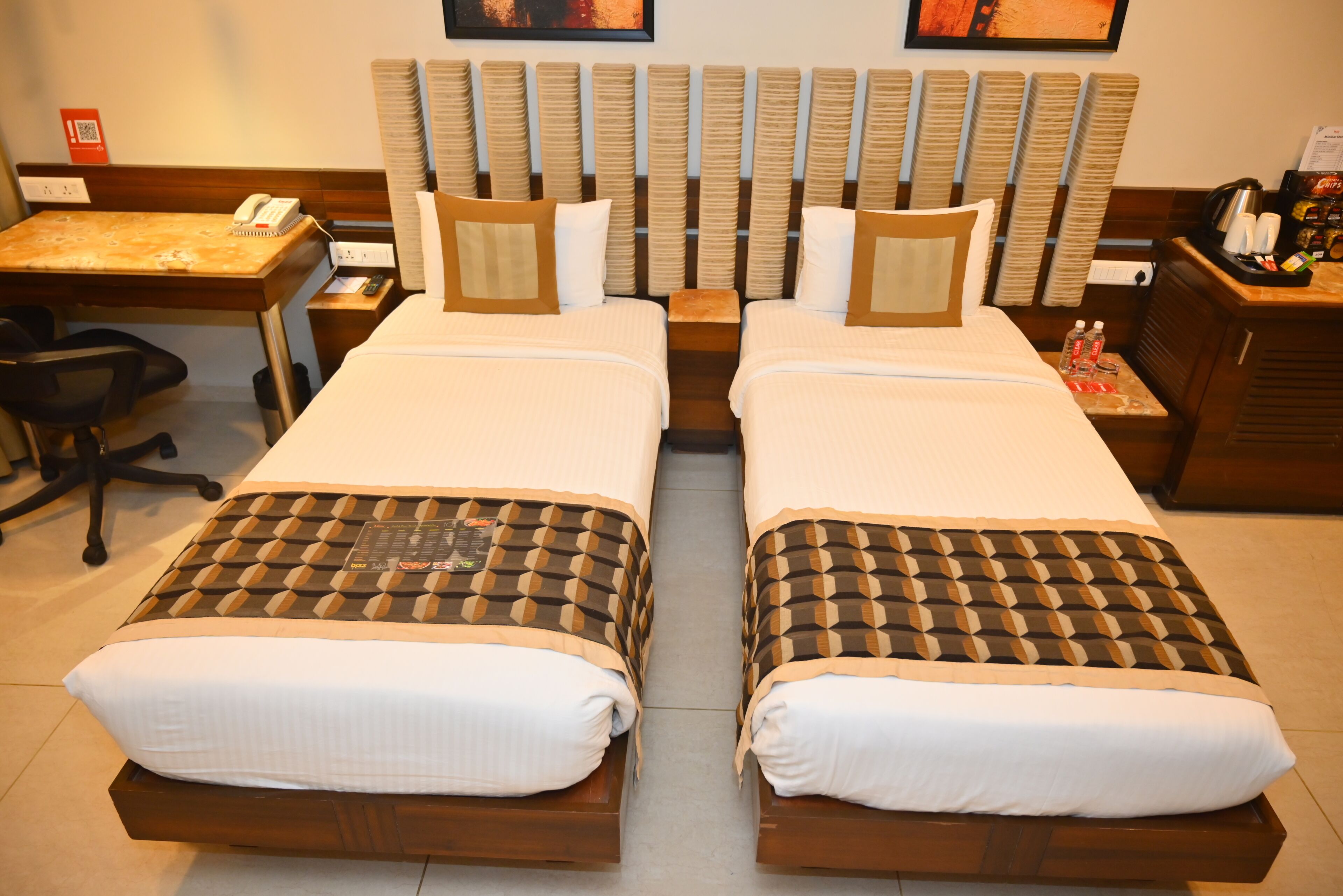 A hotel room with two beds, a desk, a chair, a radiator, and a small table with a phone and a tray of items.
