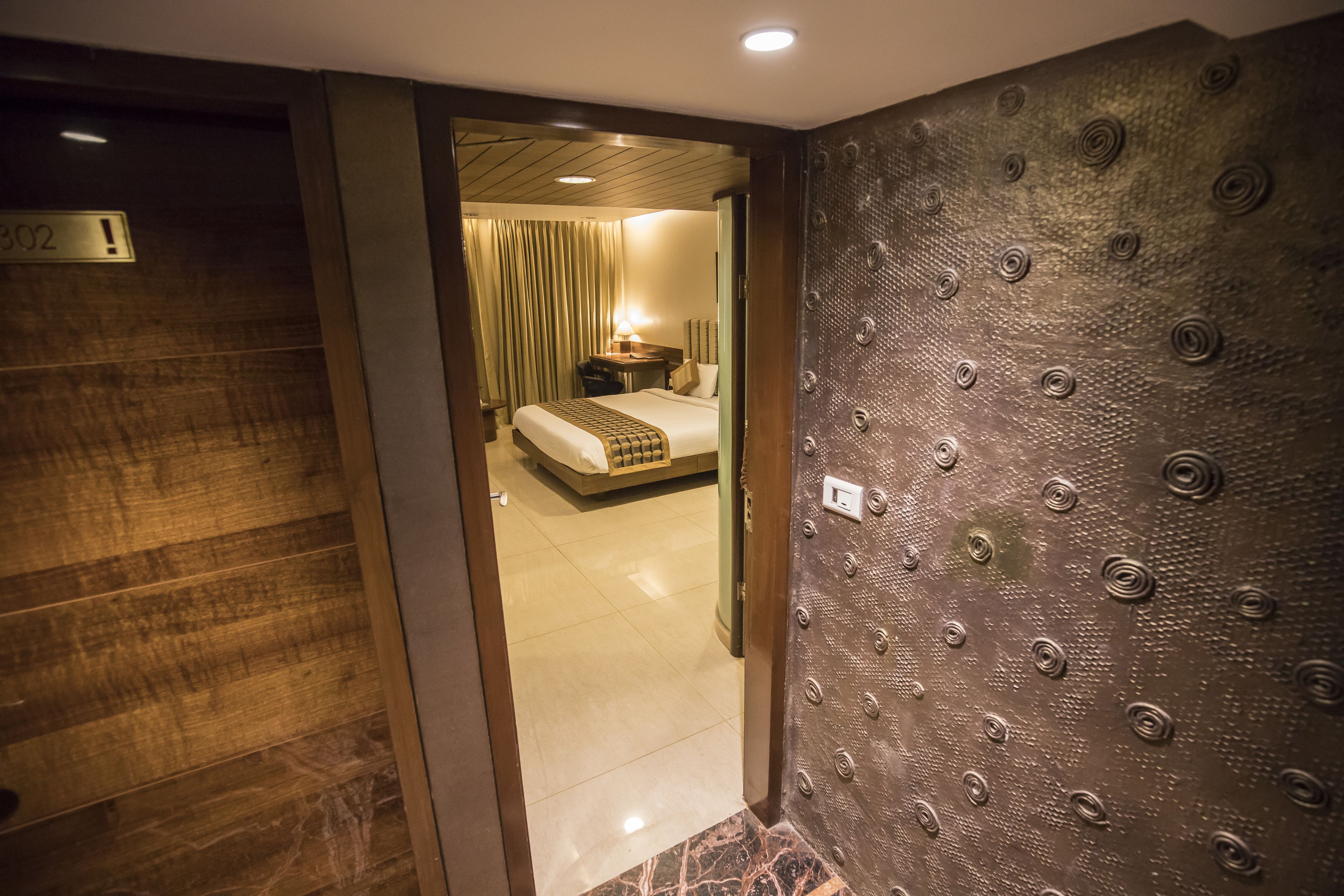 Habitación de hotel con piso de madera, cama con cabecera, mesita de noche con lámpara y pared con textura.