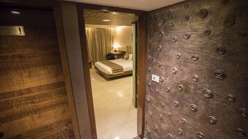 A hotel room with a wooden floor, a bed with a headboard, a bedside table with a lamp, and a textured wall.