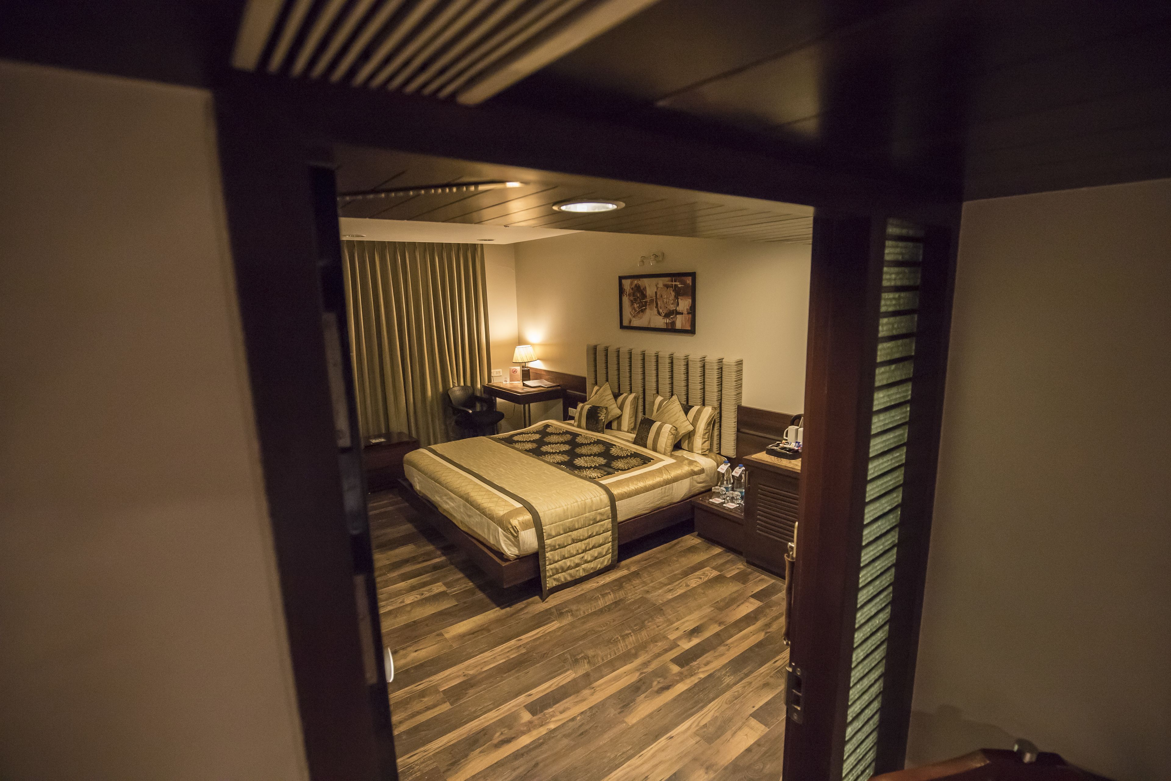 A hotel room with a large bed, a bedside table with a lamp, a headboard with decorative pillows, and a framed picture on the wall.