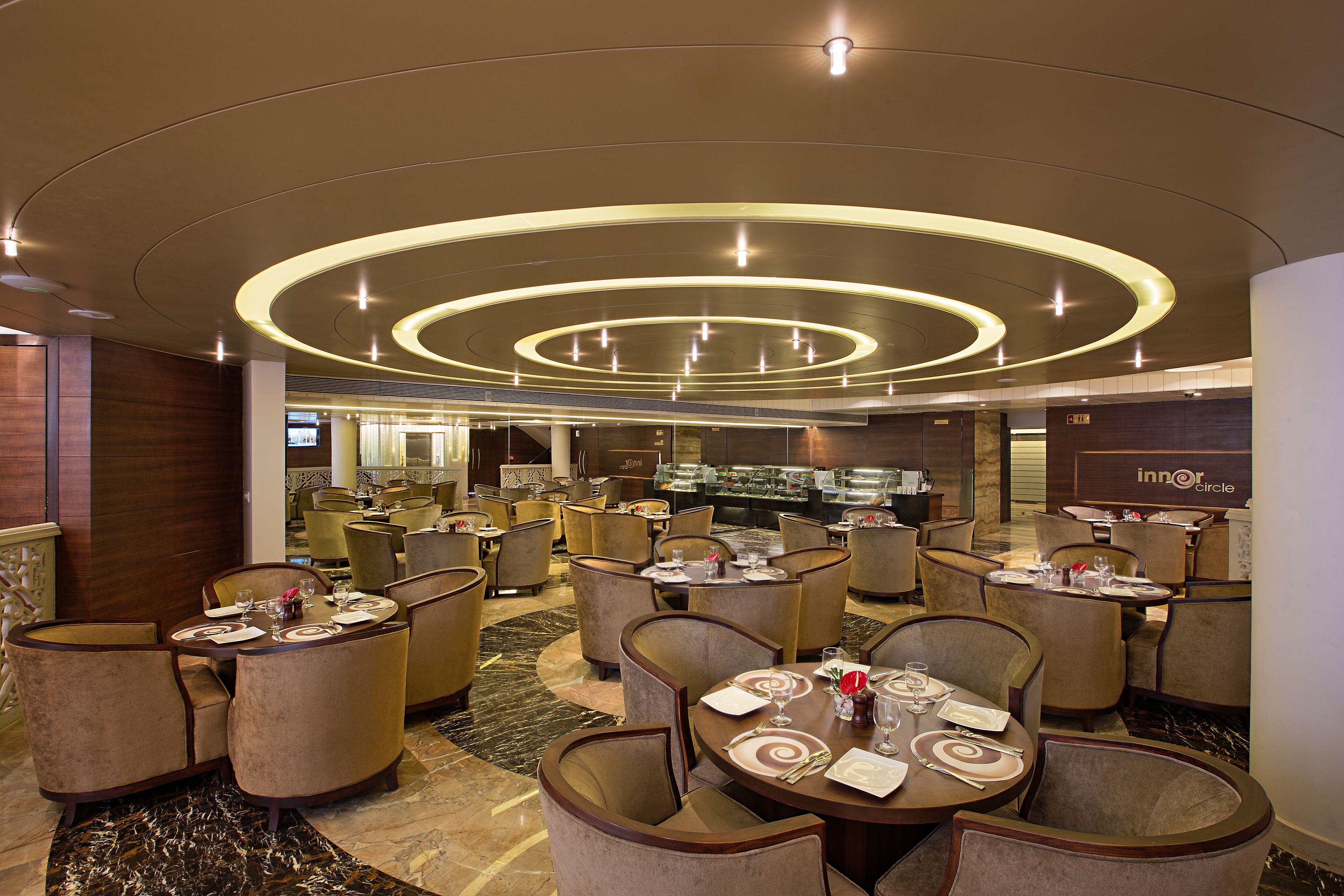 Un amplio comedor de hotel con varias mesas y sillas redondas, una barra central y un diseño de techo moderno.