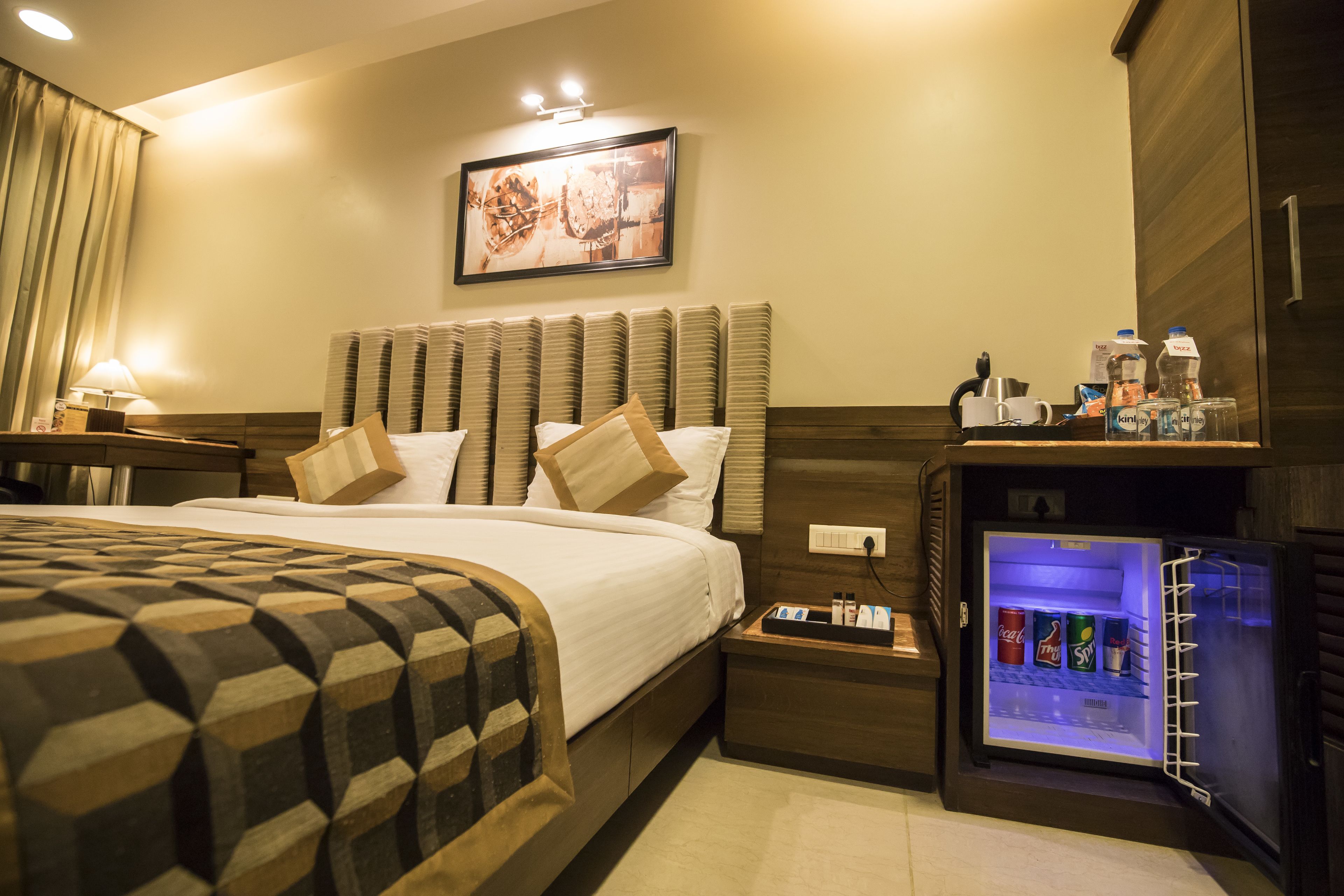 A hotel room with a bed, a nightstand, a wall-mounted refrigerator, and a painting on the wall.