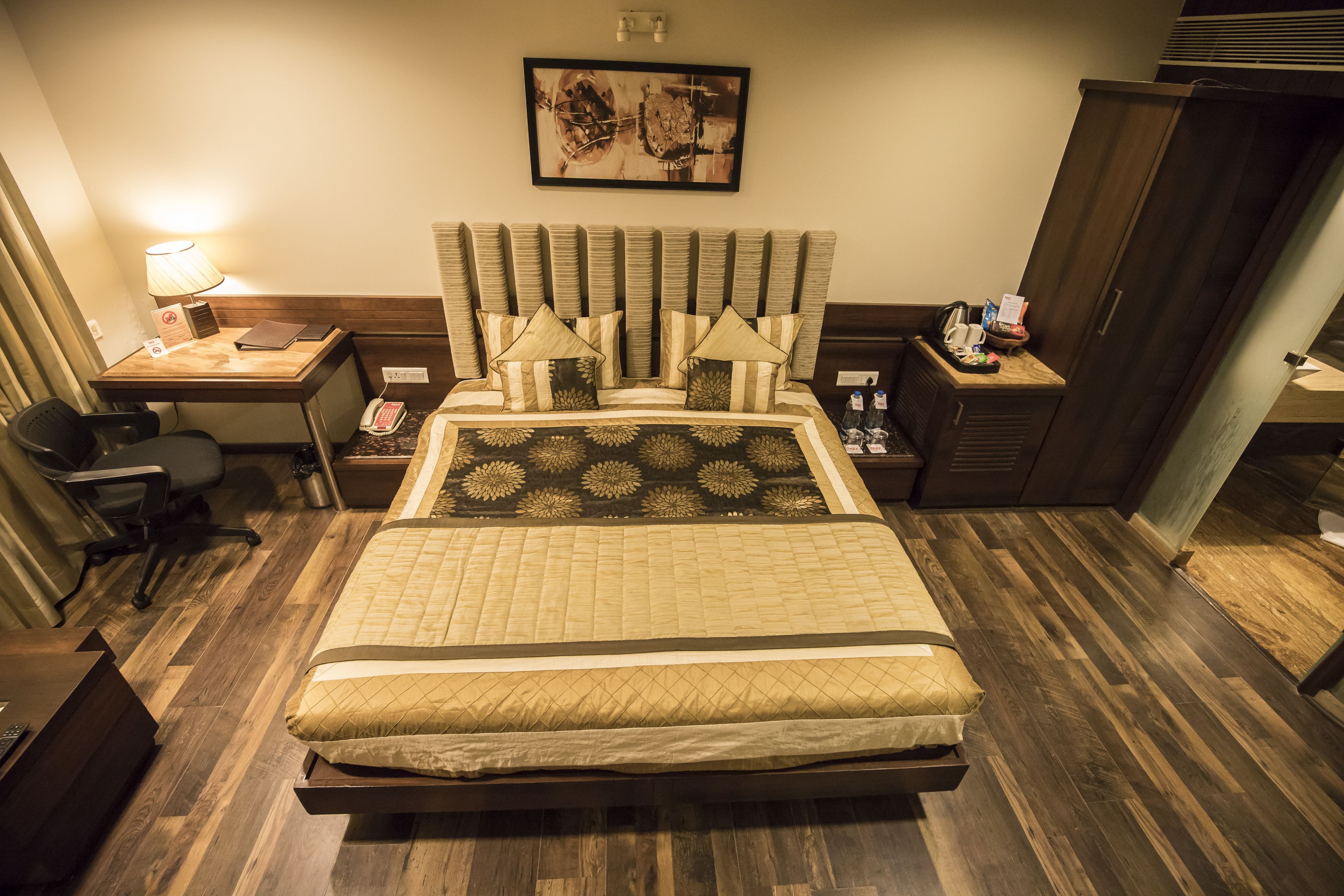 A hotel room with a large bed, a desk with a lamp, a chair, and a nightstand with a phone and a bottle.