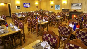 A large dining hall with wooden paneling, checkered upholstered chairs, and a chandelier. Tables are set with plates, glasses, and cutlery. A television is mounted on the wall, and a fireplace is visible in the background.