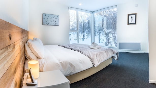 A modern bedroom with a large window, a wooden headboard, a bedside table with a lamp, and a radiator. - Ikigai (Kutchan)