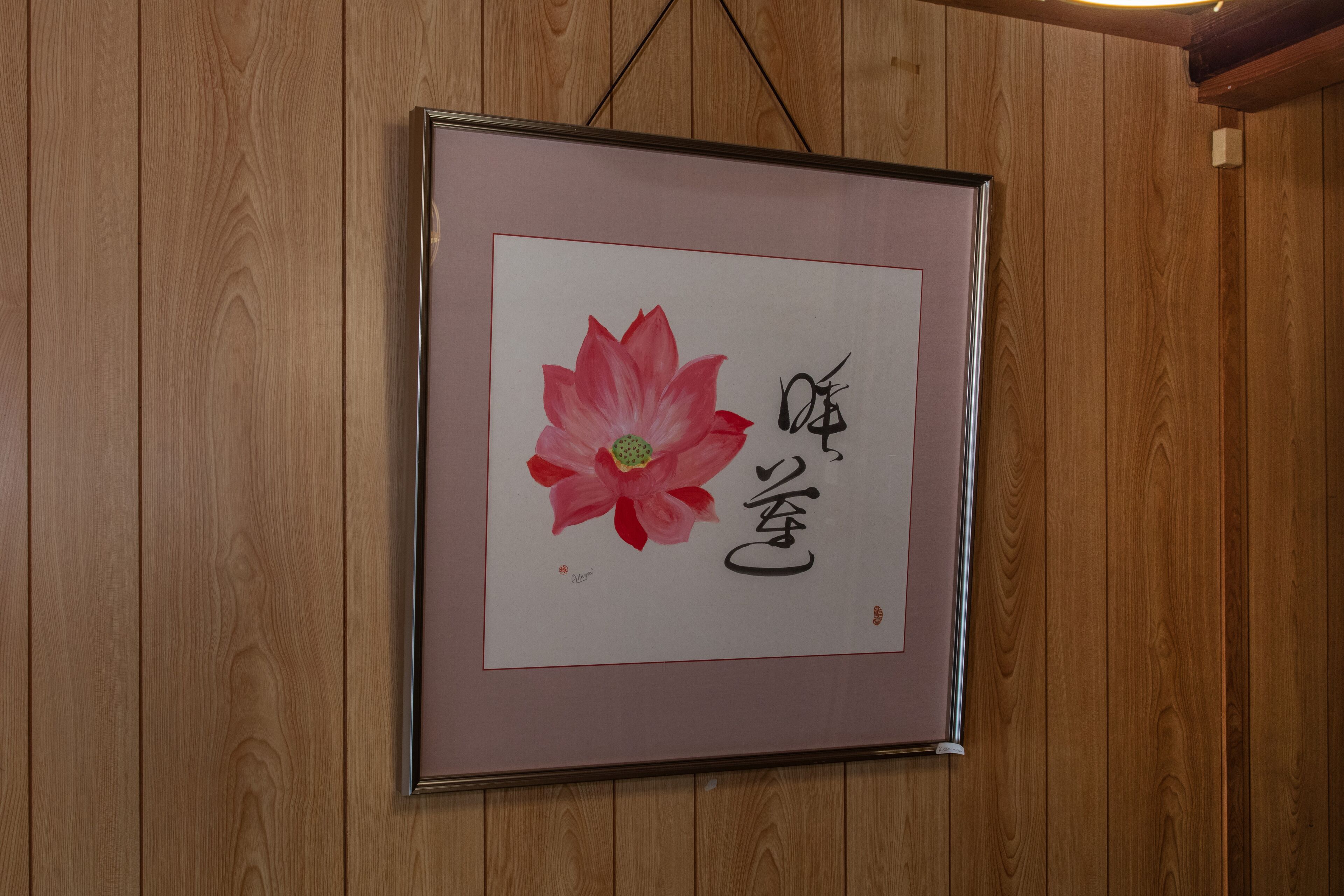 Une œuvre d’art encadrée représentant une fleur rose et des caractères japonais, le tout accroché à un mur en bois.