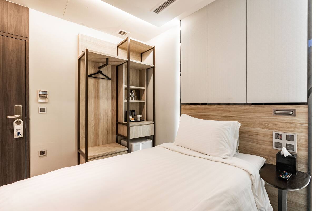 Quarto de hotel com cama, mesa de cabeceira com controle remoto, guarda-roupa e pequena escrivaninha com uma xícara e uma caixa de lenços.