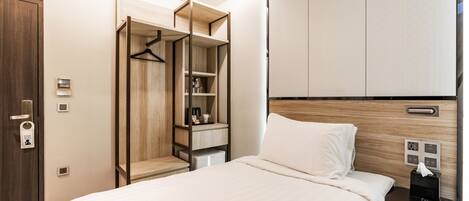 A hotel room with a bed, a bedside table with a remote control, a wardrobe, and a small desk with a cup and a tissue box.