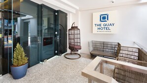 Un moderno vestíbulo de hotel con puertas de cristal, una planta en maceta, mobiliario de mimbre y un cartel de The Quay Hotel.