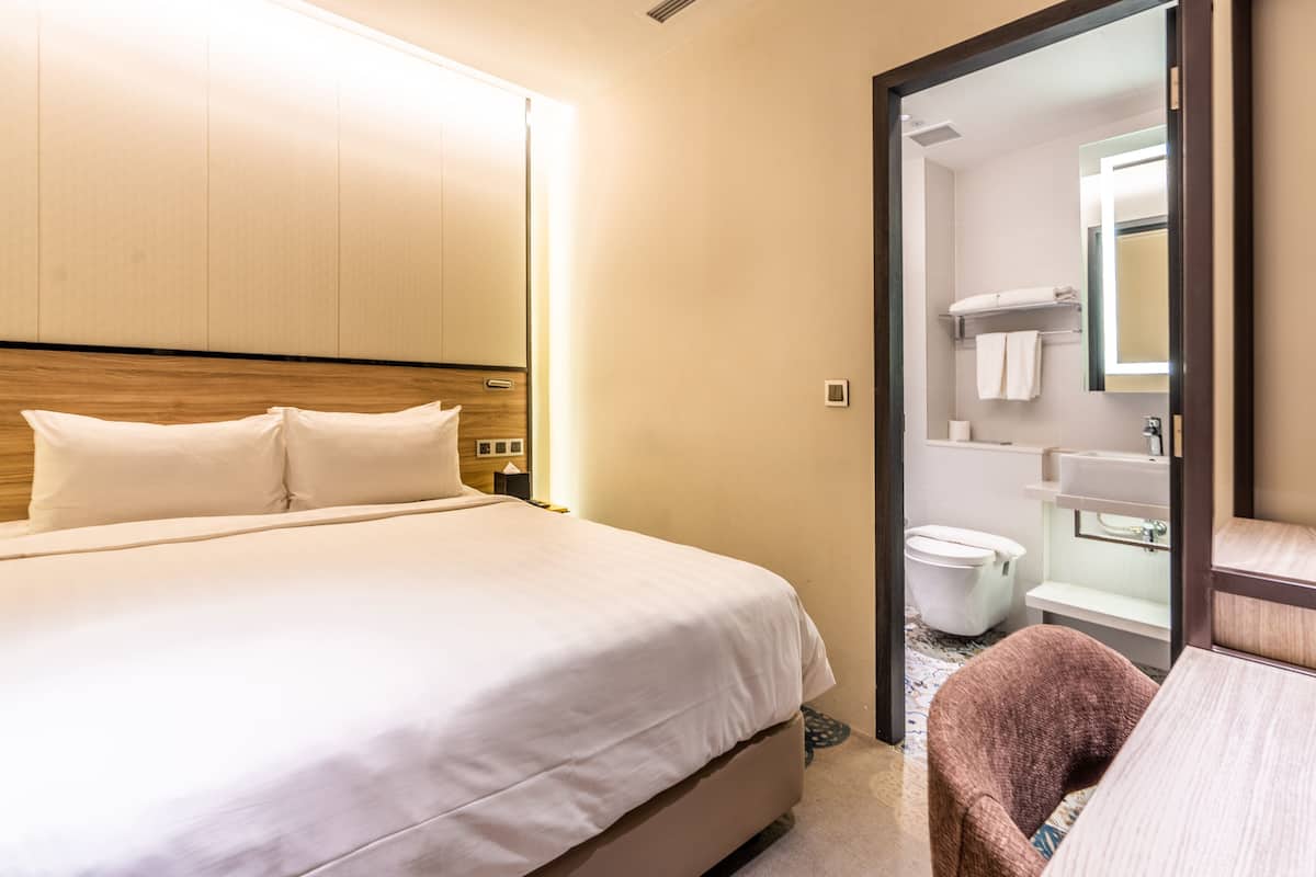 Quarto de hotel com uma cama grande, cabeceira de madeira, uma mesinha, uma cadeira e um banheiro visível por uma porta aberta.