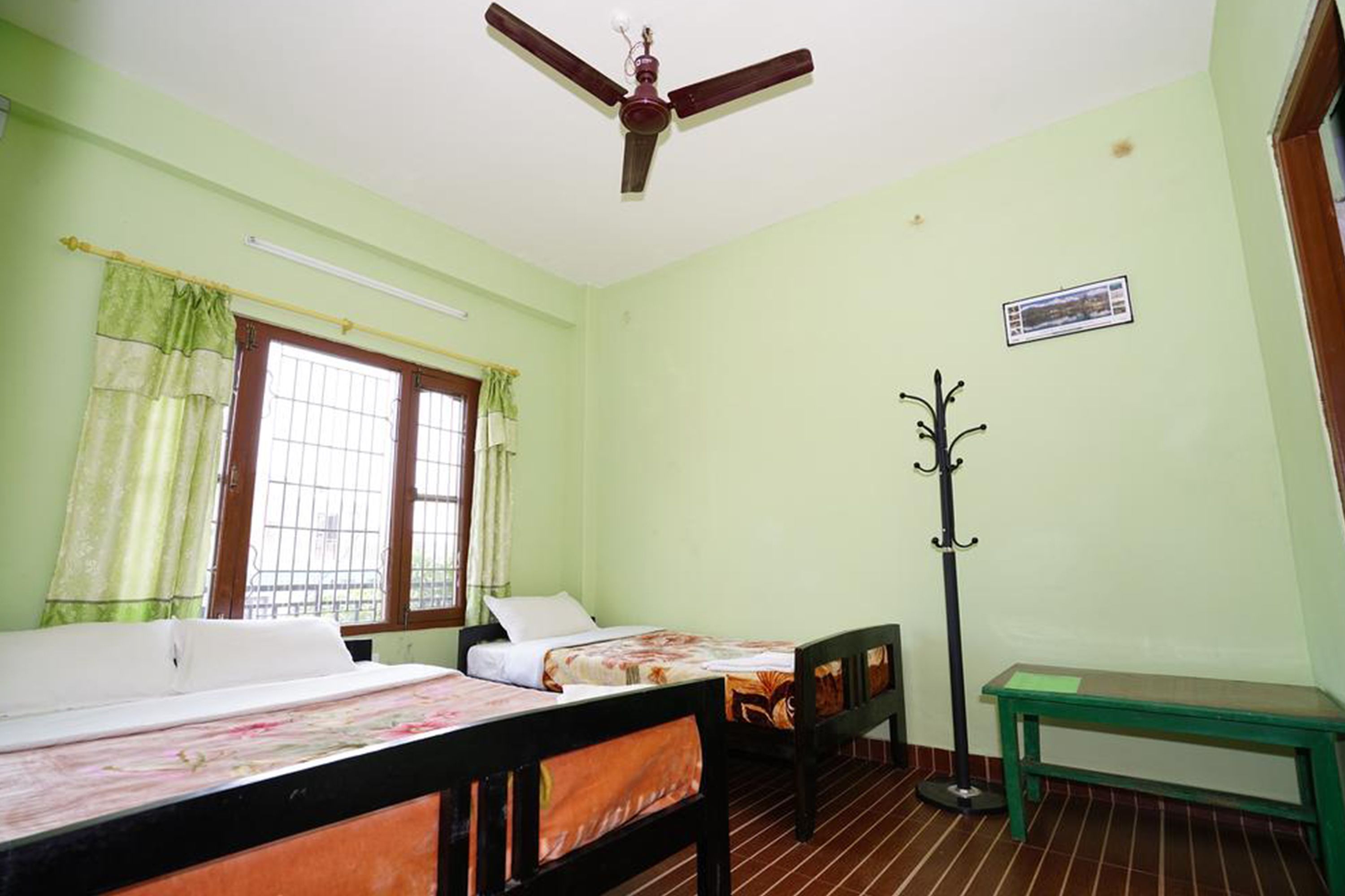 Una habitación con dos camas, ventilador de techo, ventana con cortinas, perchero y un banco.