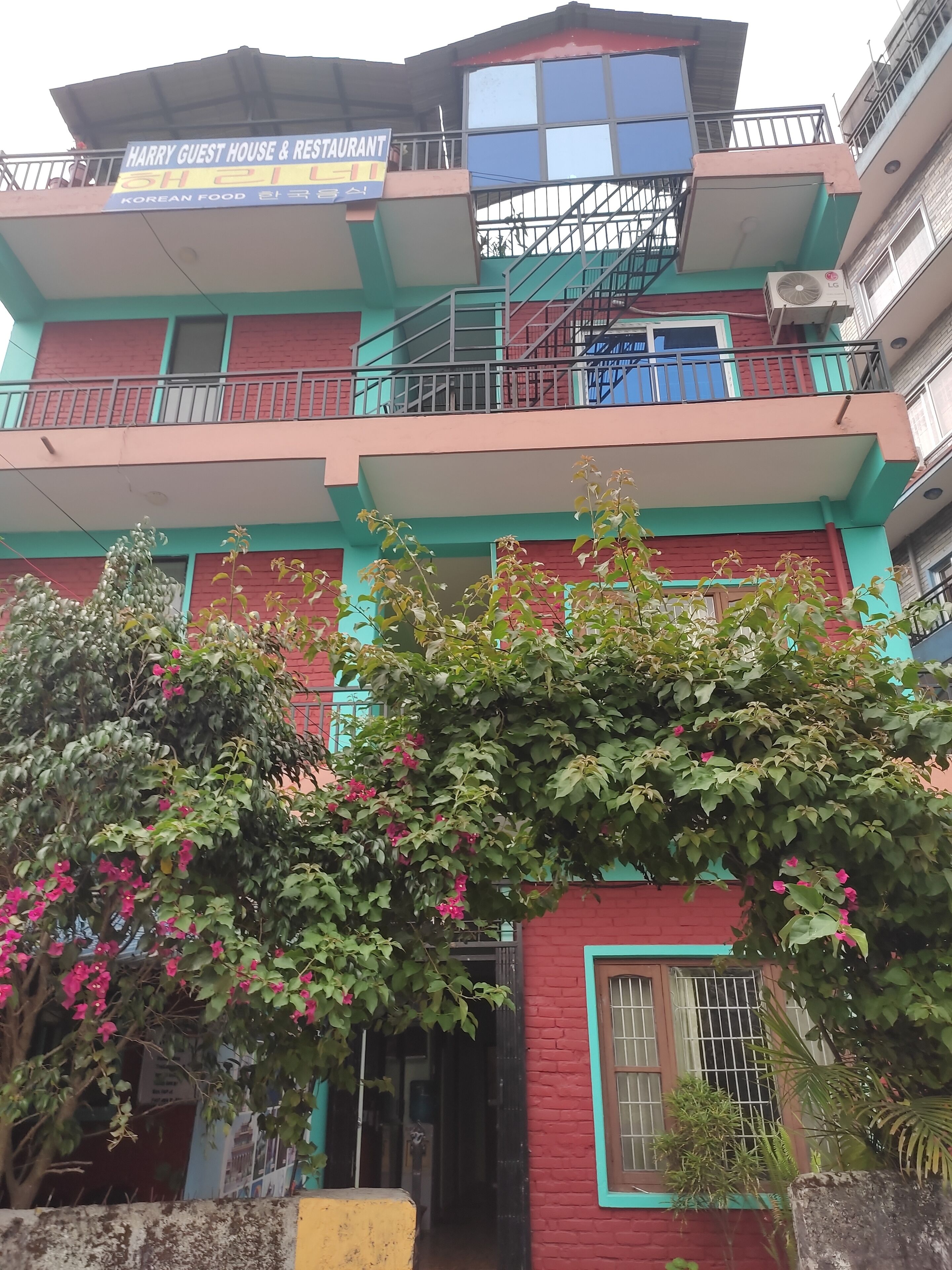 A multi-story building with balconies, a sign for a Korean restaurant, and flowering plants in front.