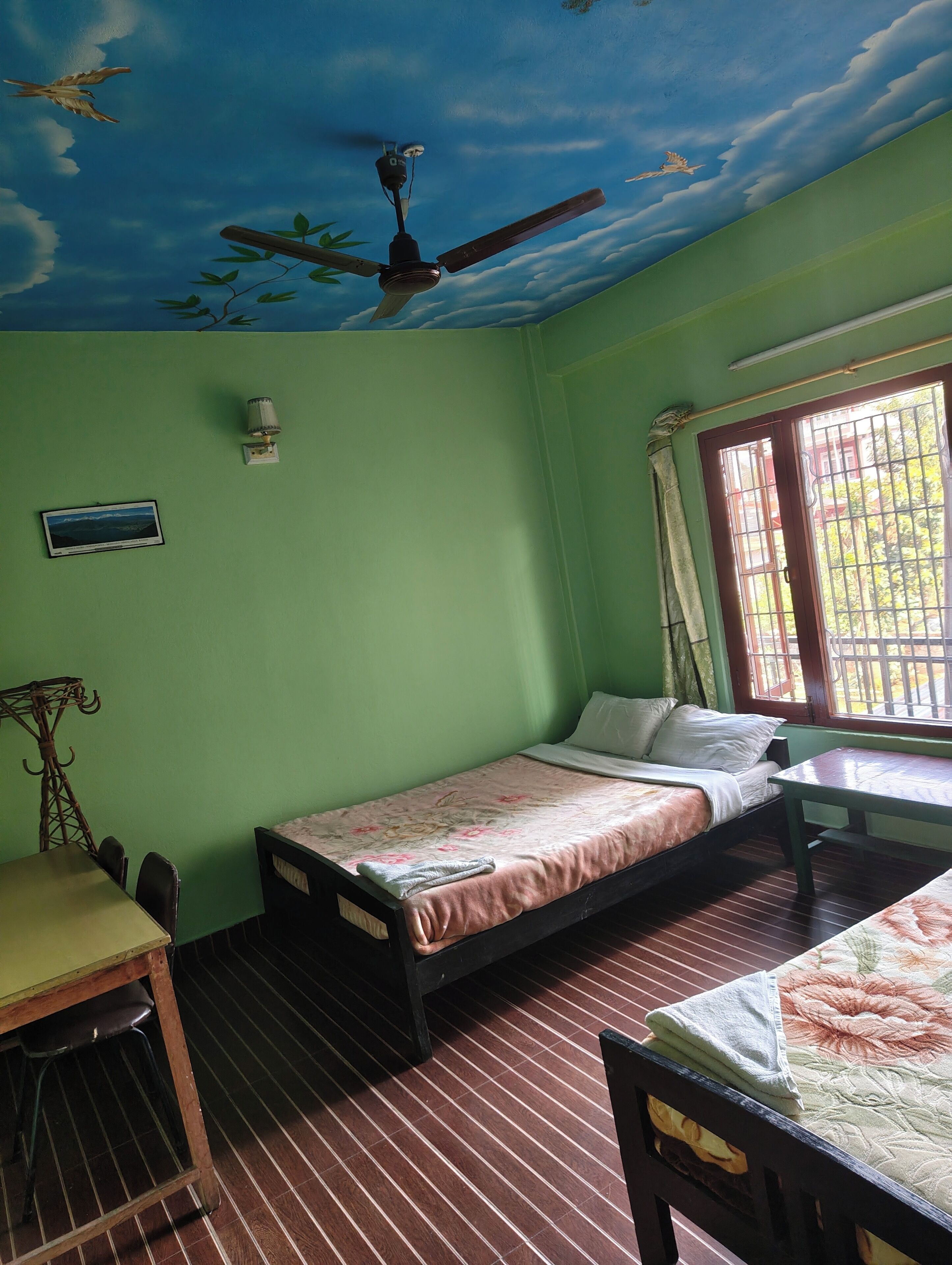 Une pièce comprenant un lit, une table, une chaise, un ventilateur de plafond, une fenêtre avec des barreaux et une lampe fixée au mur.