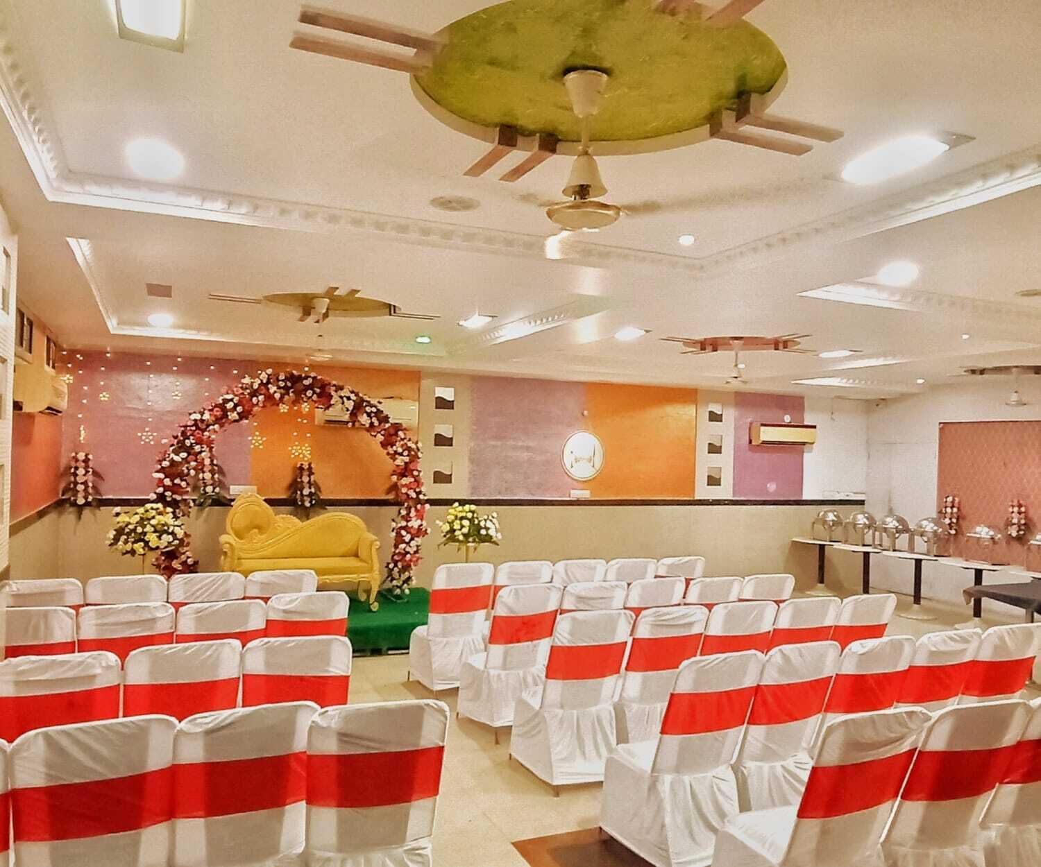 Ein Festsaal mit Stuhlreihen, einer Bühne mit floralen Dekorationen und Deckenventilatoren.