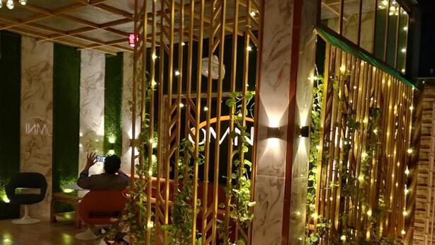 A modern, well-lit outdoor seating area with a glass enclosure, potted plants, and a wooden fence. - Omni Plaza (Jodhpur)