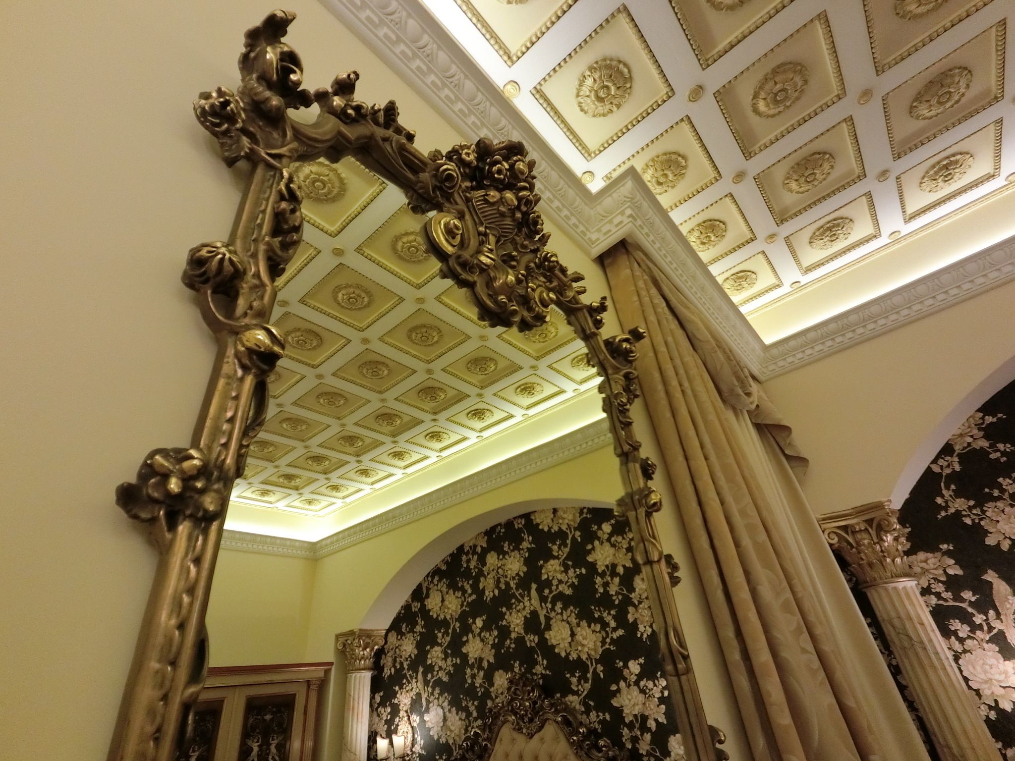 An ornate mirror with a floral design, a ceiling with decorative panels, and a wall with a floral wallpaper pattern.