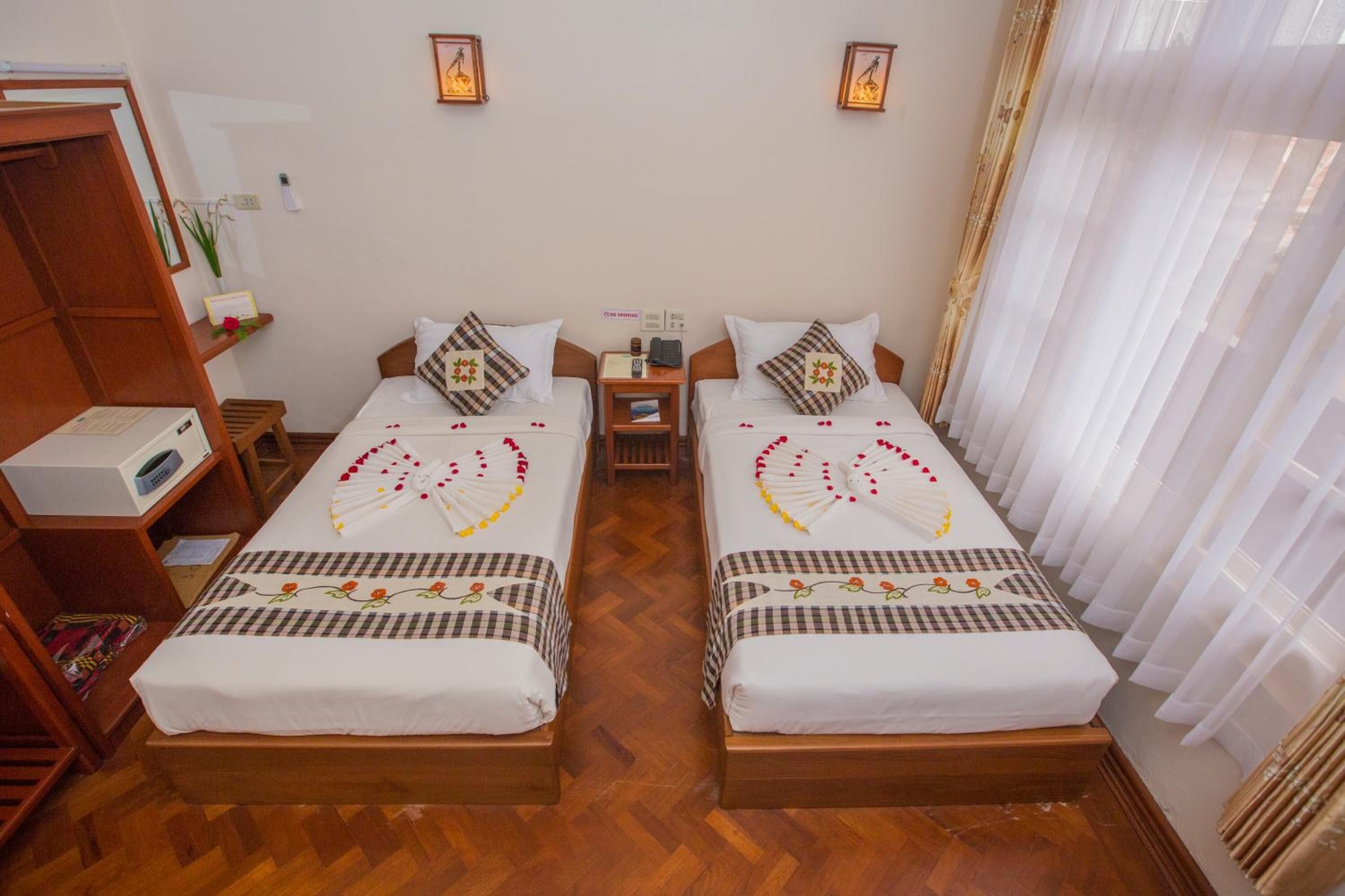 A hotel room with two beds, wooden flooring, a small bedside table with a lamp, and a mirror on the left wall.