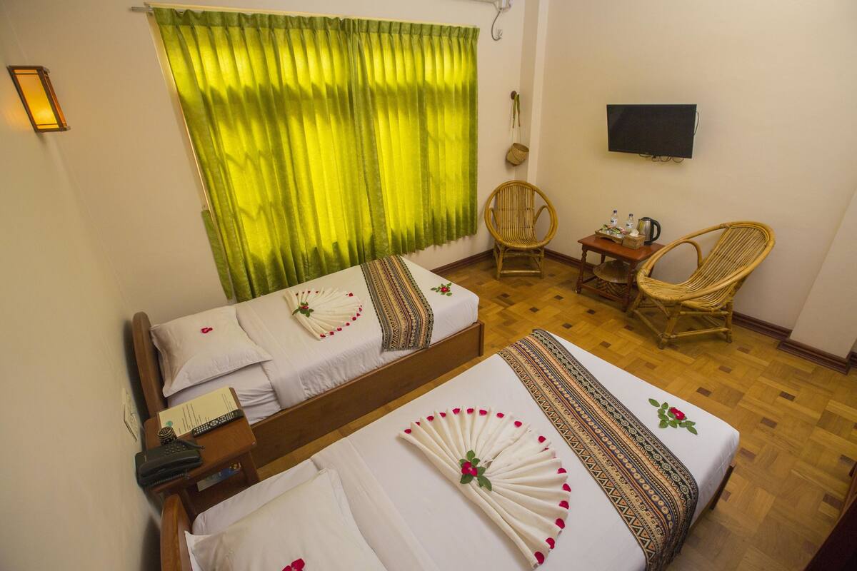 A hotel room with two beds, a television, a wicker chair, and a small table with a kettle and bottles.