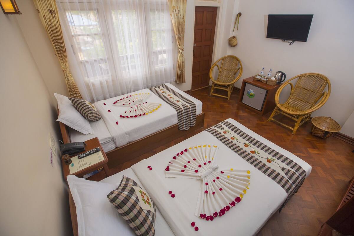 A hotel room with two beds, a TV, wicker chairs, and a small table with a kettle.