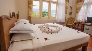 A neatly made bed with rose petals and a heart-shaped arrangement, a wicker chair, a small table with a kettle, and a view of greenery through the window. - Inle Apex Hotel (Nyaungshwe)