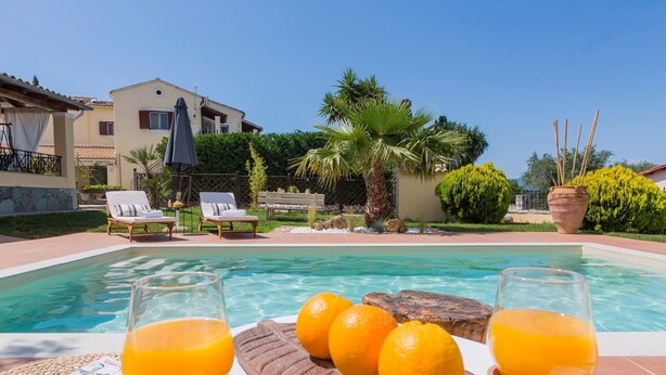 Een zwembadgedeelte met ligbedden, een houten tafel met sinaasappels en glazen jus d'orange, en een gebouw op de achtergrond.