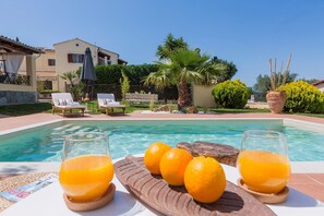A poolside area with lounge chairs, a wooden table with oranges and glasses of orange juice, and a building in the background. - Villa Caramel (Corfu)
