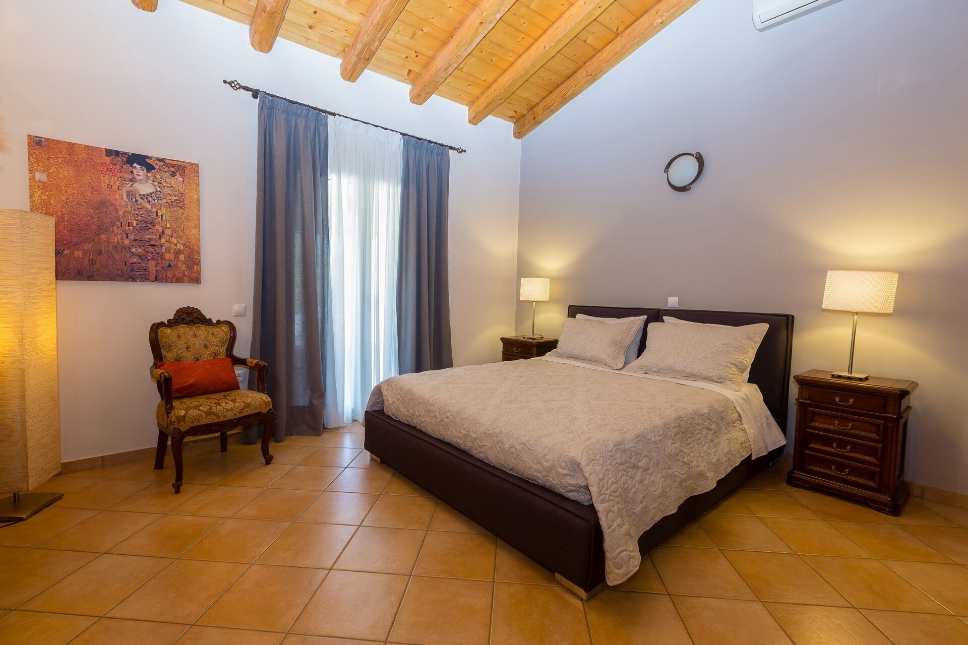 A bedroom with a large bed, a chair, a painting, and a clock on the wall.