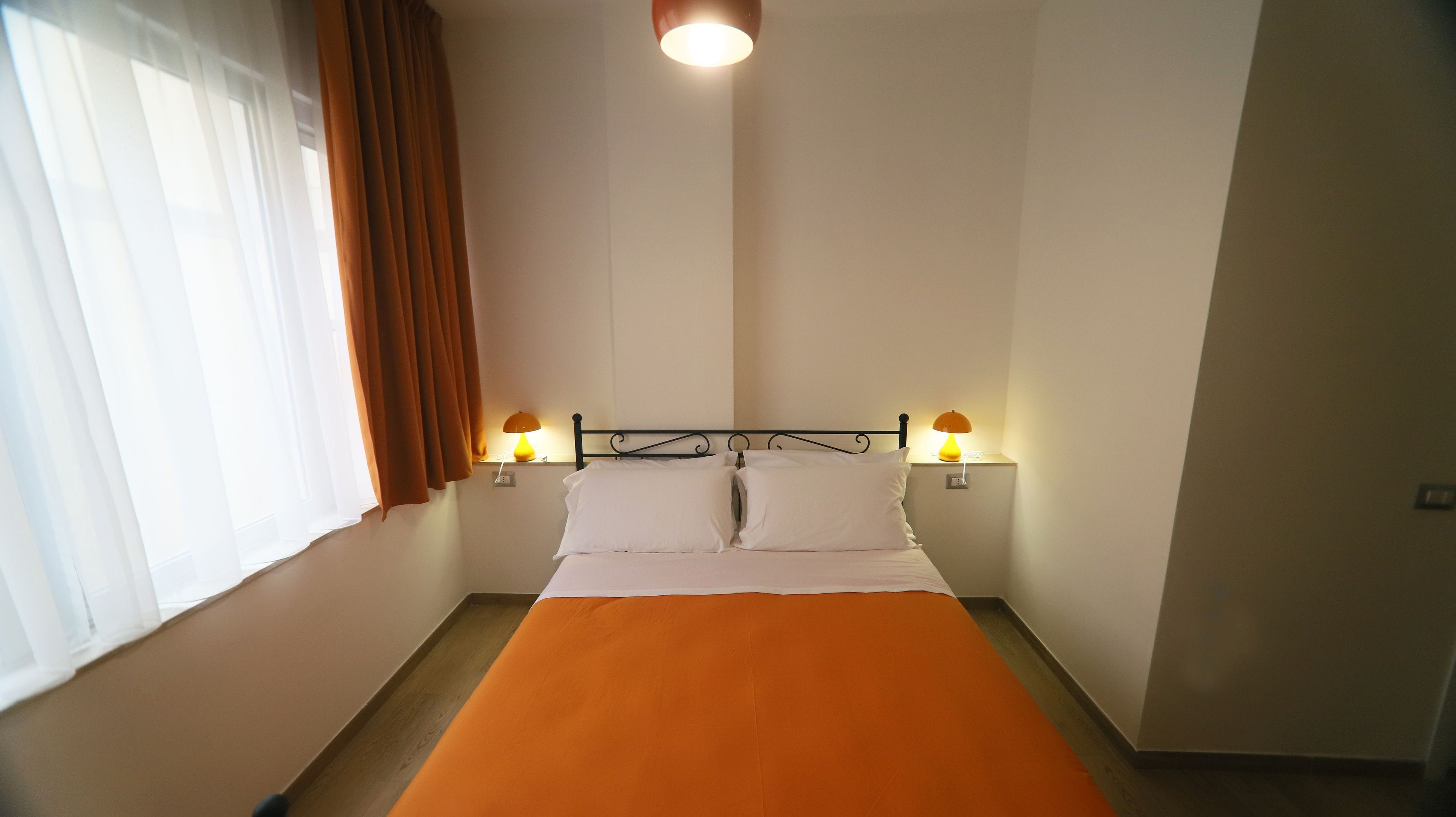 A single bed with an orange bedspread, two bedside lamps, and a window with brown curtains.