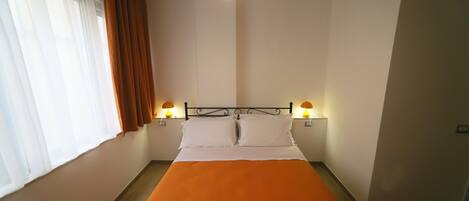A single bed with an orange bedspread, two bedside lamps, and a window with brown curtains.