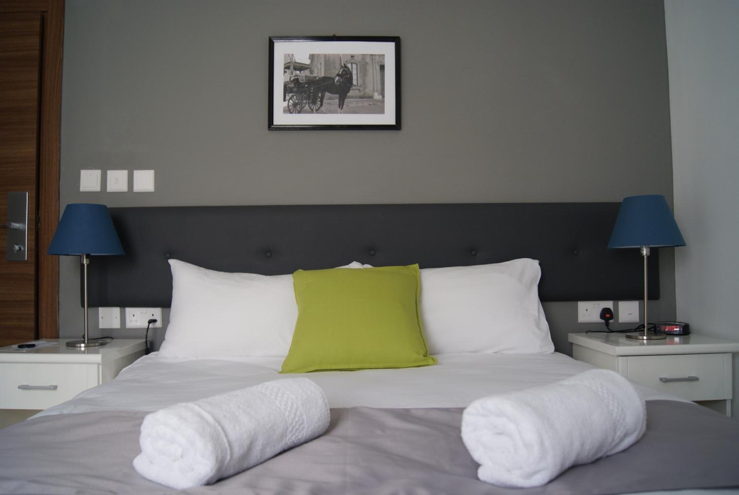 A neatly made bed with white and gray linens, two bedside lamps, a green pillow, and a framed picture on the wall.