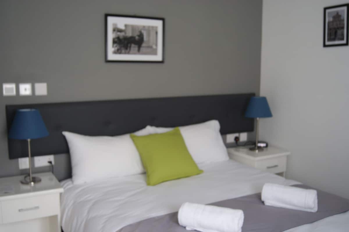 Habitación de hotel con cama, dos lámparas de noche, mesita de noche, un cuadro enmarcado en la pared y una toalla sobre la cama.