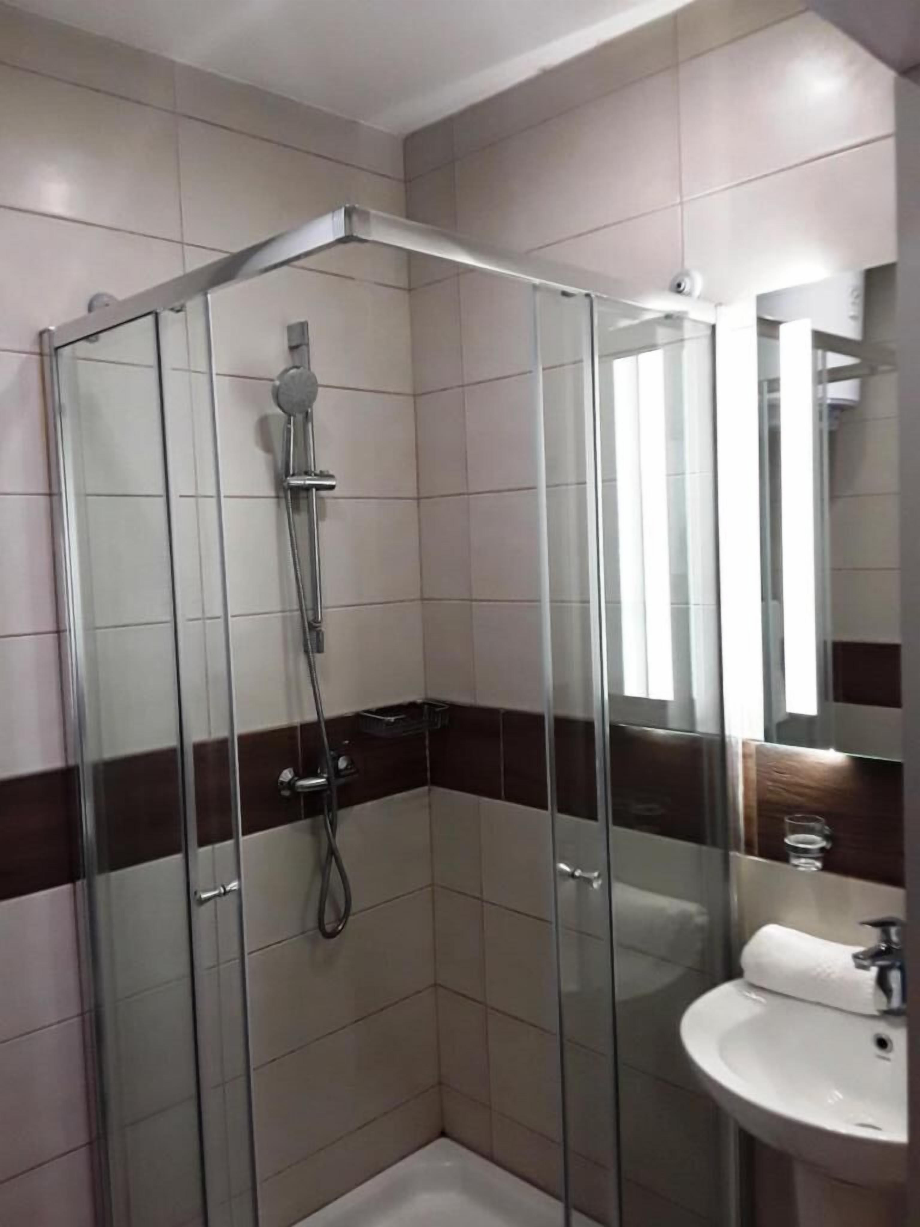 Ducha con mampara de vidrio, cabezal de ducha manual, lavamanos blanco con espejo y detalle de borde en cerámica marrón oscuro.