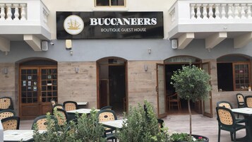 A building named 'BUCCANEERS Boutique Guest House' with outdoor seating and a small tree in the foreground.