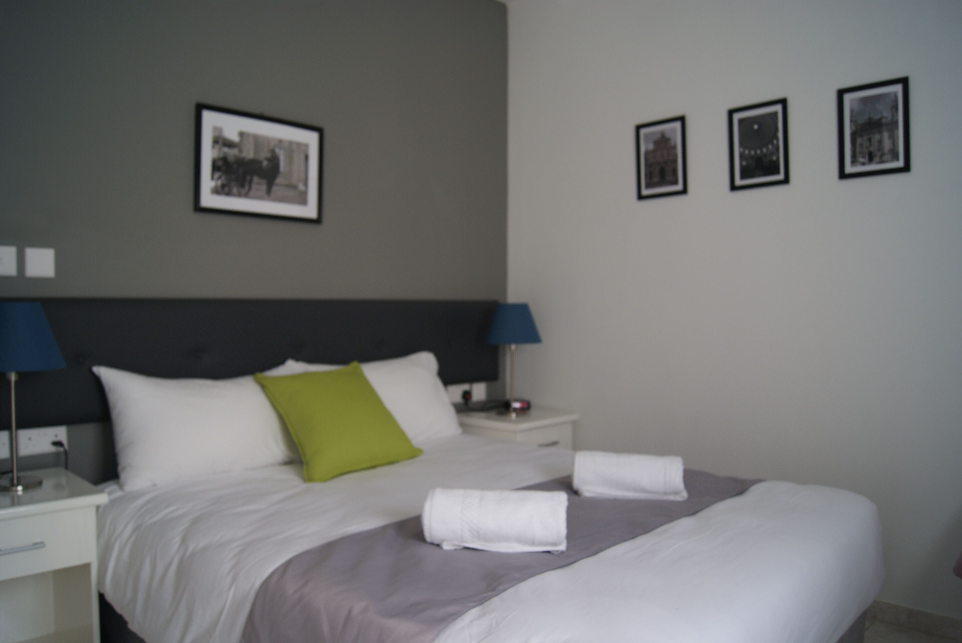 A neatly made bed with white and grey bedding, two bedside lamps, and a nightstand with a lamp. There are three framed pictures on the wall.