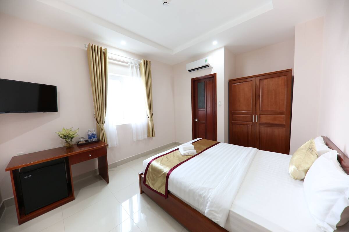 A hotel room with a bed, a desk with a television, a wardrobe, and an air conditioning unit.