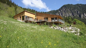 Una casa moderna de madera con balcón, ubicada en una ladera con montañas al fondo.