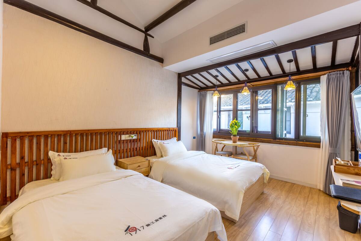 Een hotelkamer met twee bedden, houten hoofdborden, een kleine tafel met een plant en een raam met gordijnen.