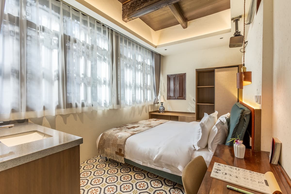 Een hotelkamer met een bed, een bureau met een lamp, een klein wastafeltje en een vloer met een patroon.