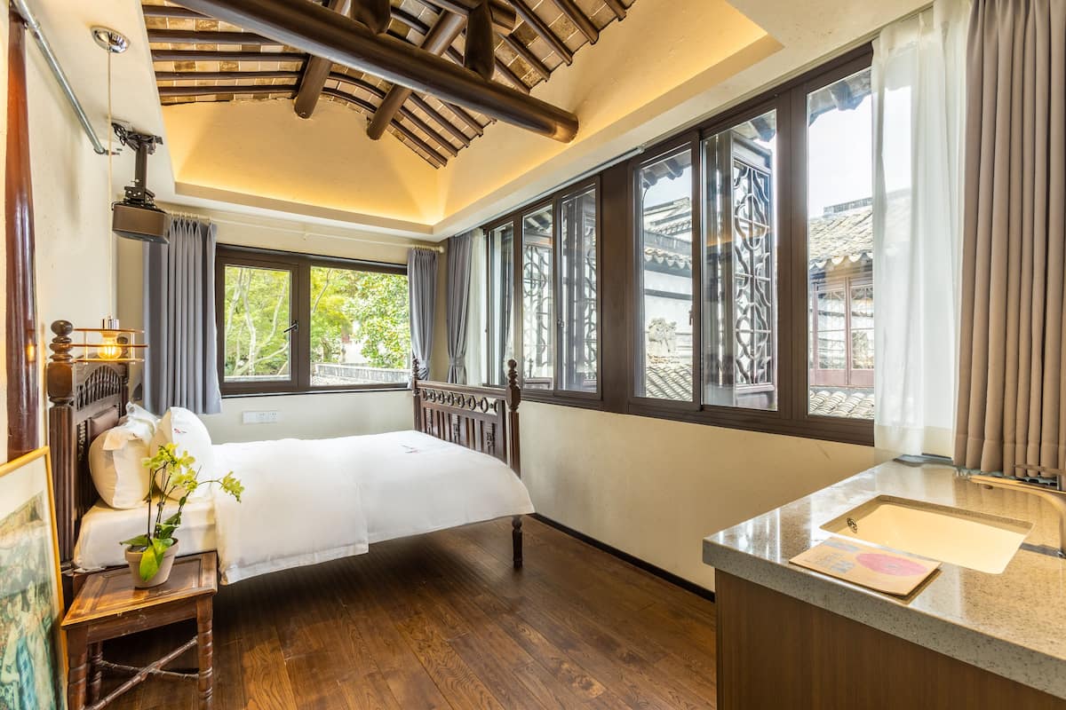 Een slaapkamer met een houten bed, een nachtkastje met een lamp, een groot raam met gordijnen en uitzicht op een traditioneel gebouw.