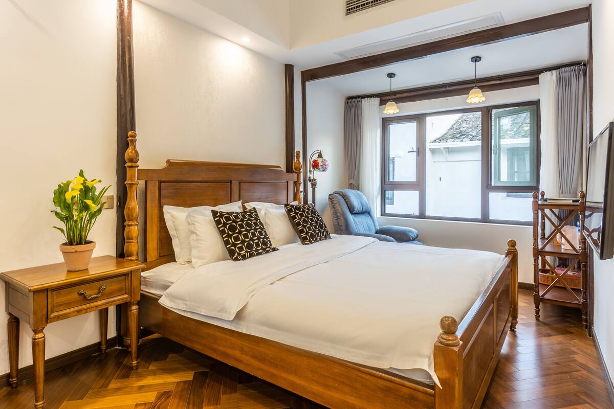 Een houten bed met witte beddengoed, een nachtkastje met een plant, een stoel en een raam met gordijnen.